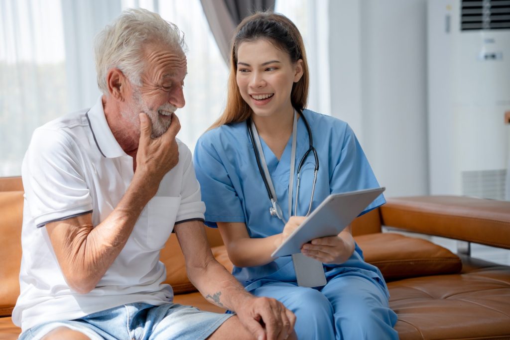 An older adult man in a light-colored button-down shirt and shorts sits next to a younger medical provider wearing light blue scrubs. He is looking at the depression treatment plan on a clipboard she is holding in front of him while she smiles at him. 