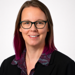 Anne Nelson, a white woman with straight dark red hair wearing glasses and a black shirt.